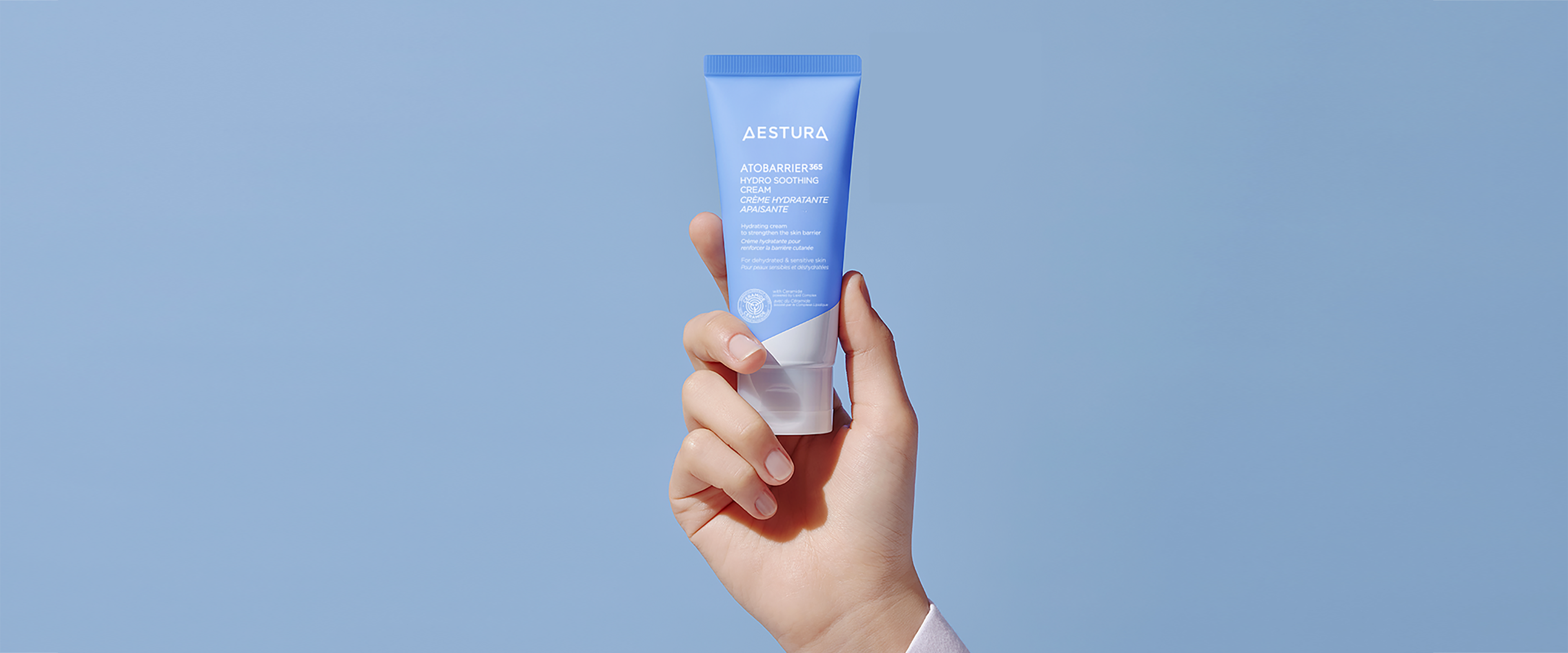 Hand holding Aestura Atobarrier365 Hydro Soothing Cream in front of blue background